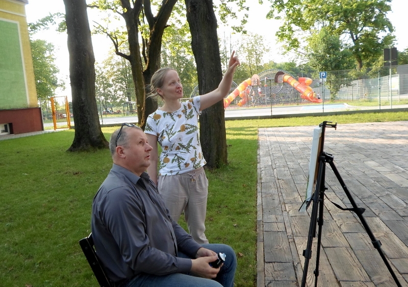 przed sztalugą na krześle stoi mężczyzna, obok niego stoi kobieta z uniesioną jedną ręką