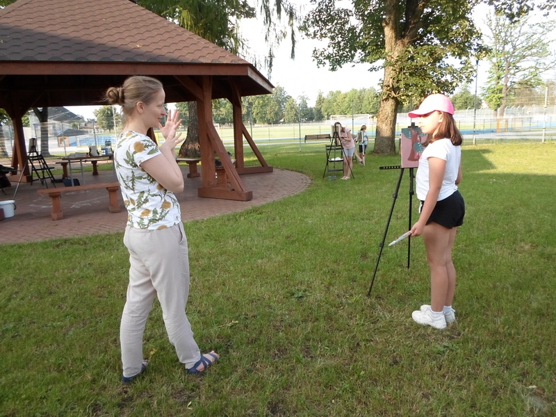 dziewczynka stoi przed sztalugą i maluje, obok niej stoi kobieta