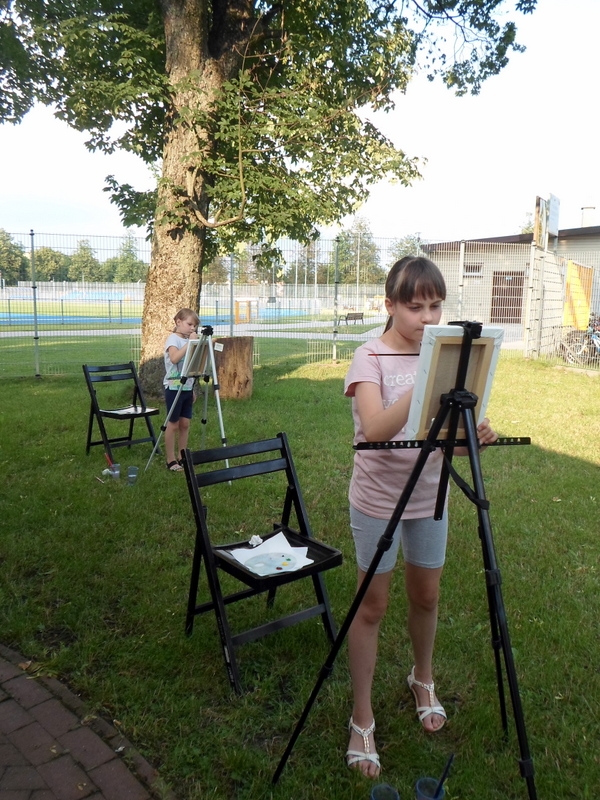 młoda uczestniczka pleneru stoi przed sztalugą i maluje obraz, za nią stoi druga dziewczynka
