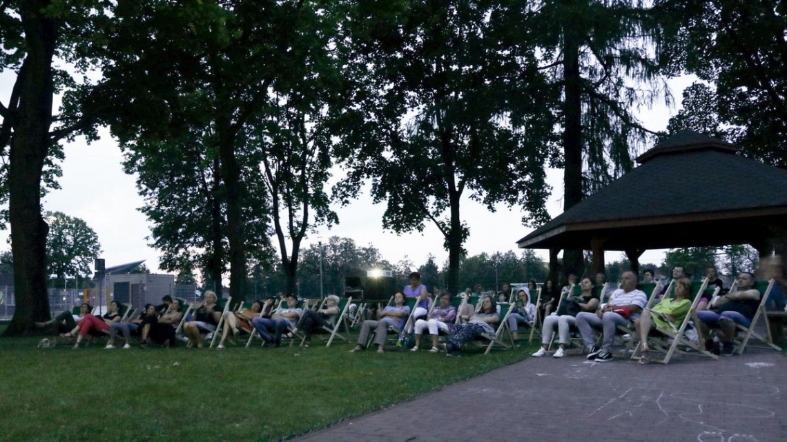 Ludzie siedzą na krzesłach, za nimi stoi wiata