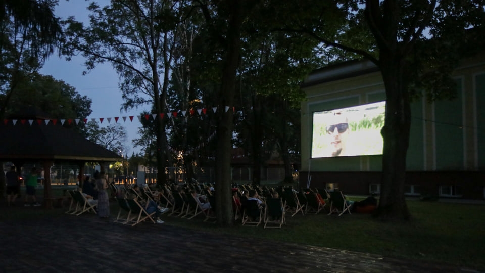 Ludzie siedzą na krzesłach, po prawej stronie ekran filmowy