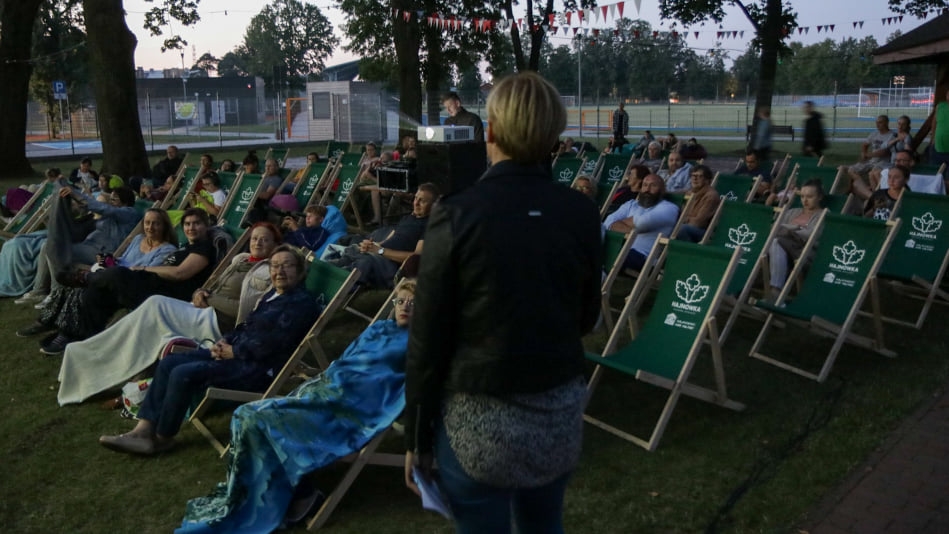 Ludzie siedzą na krzesłach, po środku stoi odwrócona tyłem kobieta