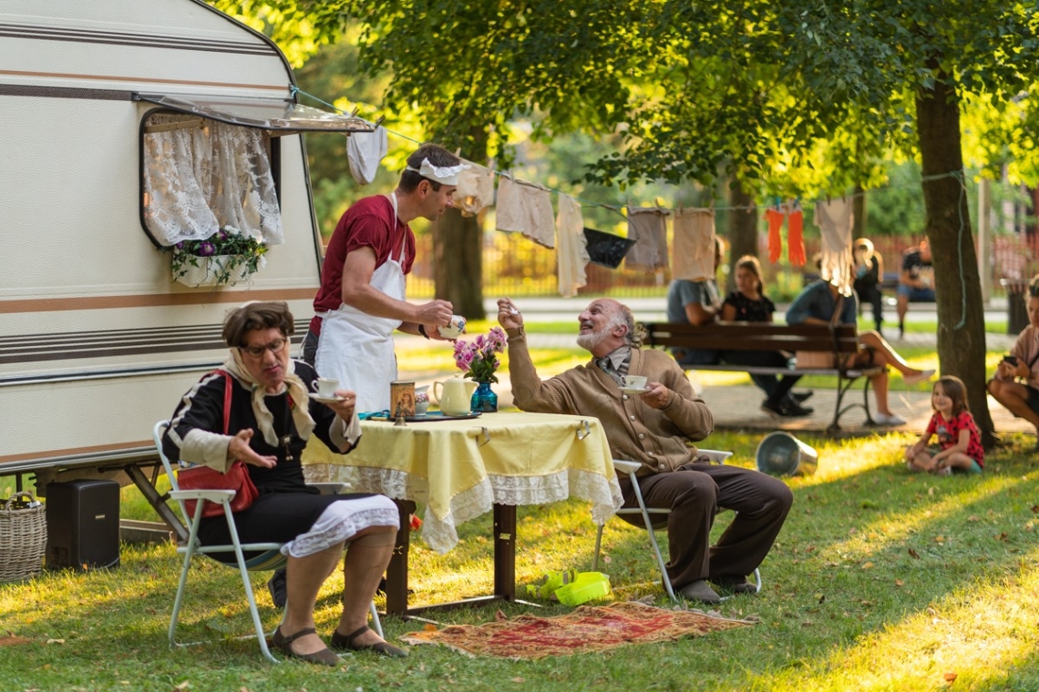 w letniej scenerii, na zielonej trawie wokół zielone drzewa, scenka aktorska. Z tyłu przycepa kempingowa, przed nią przy nirwielkim stoliku, siedzi mężczyzna i kobieta, przed stolikiem stoi mężczyna w białym fartuchu kelnera