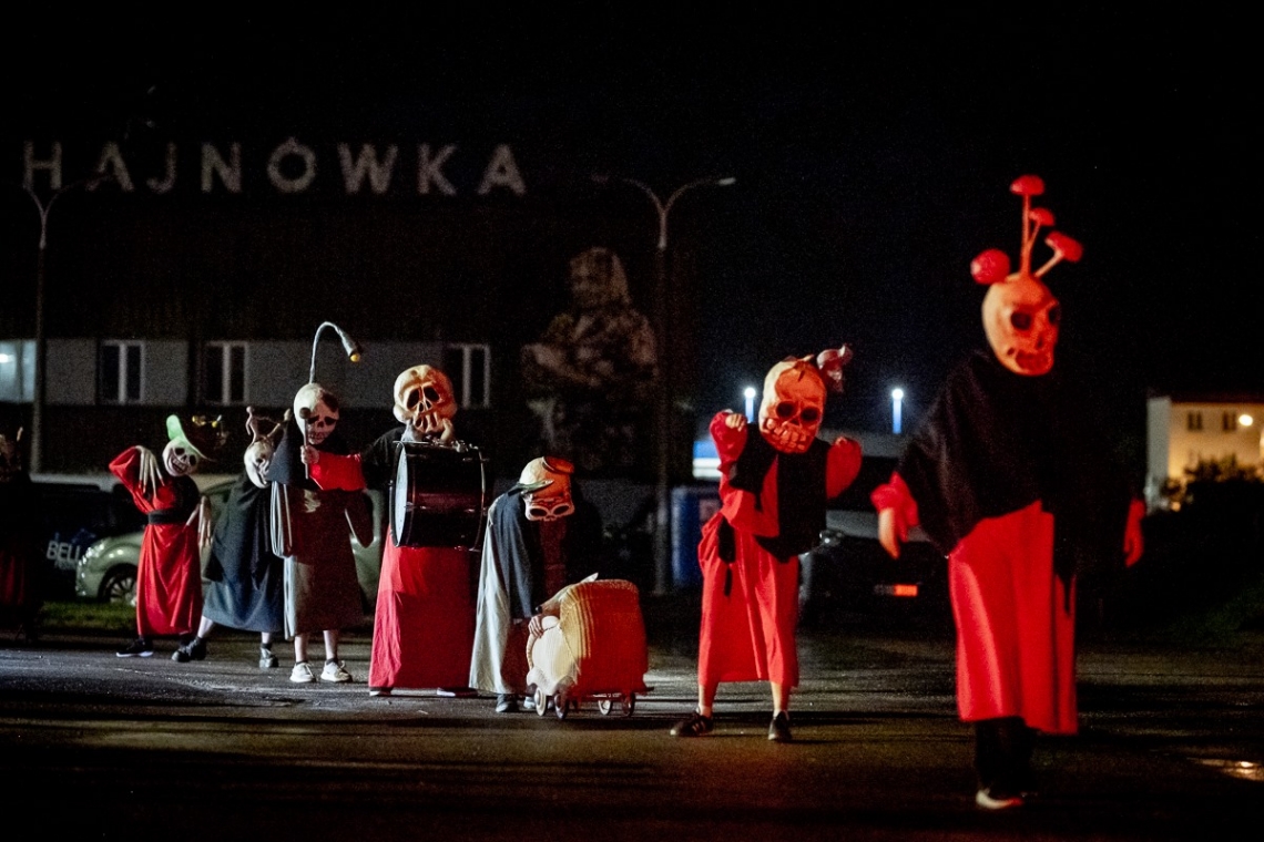 w nocnej scenerii grupa przebranych aktorów - na głowach mają białe, duże maski, u góry czarne bluzy, u dołu czerwone spódnice