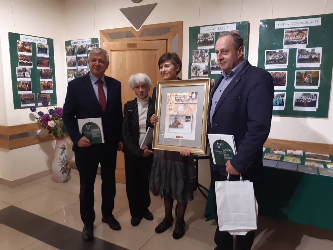 cztery osoby dwie kobiety, dwóch mężczyzn pozujacy z zdjęcia, jedna z kobiet trzyma w ręku ramkę z kartką pocztową