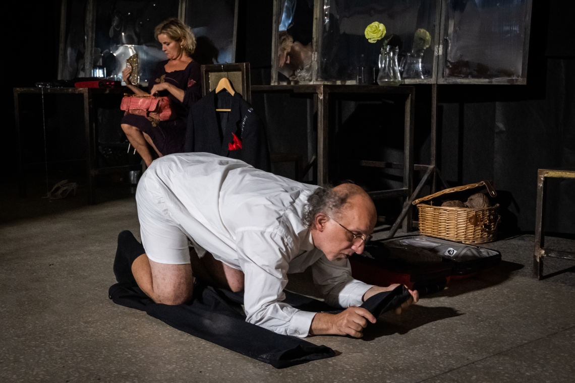 kadr z próby do spektaklu; meżczyzna w klęku podpartym na podłodze, w oddali siedząca na krześle kobieta