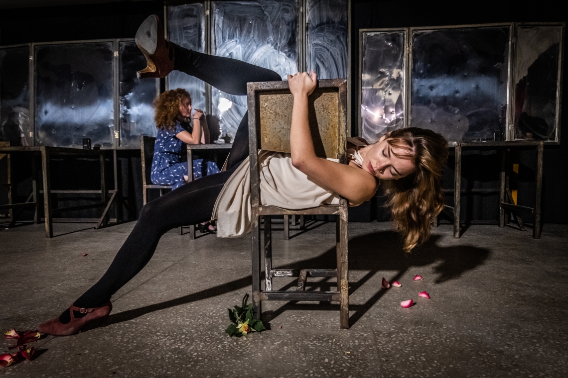 kadr z próby do spektaklu; kobieta leżąca na krześle z uniesioną jedną nogą, w oddali druga kobieta w niebieskiej sukience siedząca na krześle