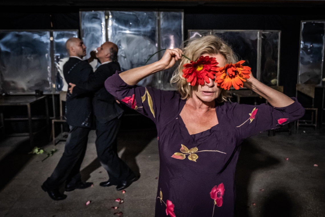 kadr z próby do spektaklu; w oddali dwoje mężczyzn tańczących w parze, na pierwszym planie kobieta, trzymajaca w rękach na wysokości oczu pomarańczowe kwiaty