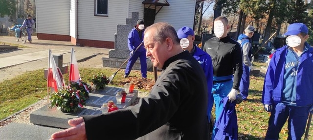 Proboszcz wskazuje ręką na groby. W tel widać mężczyzn i pomnikki