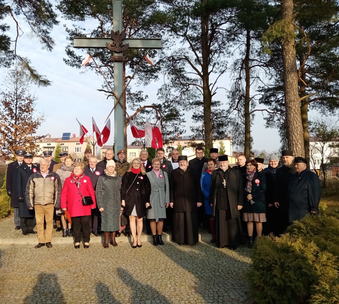 Zdjęcie grupowe samorządowców, duchownych i władz mundutowych, za nimi wysoki metalowy krzyż