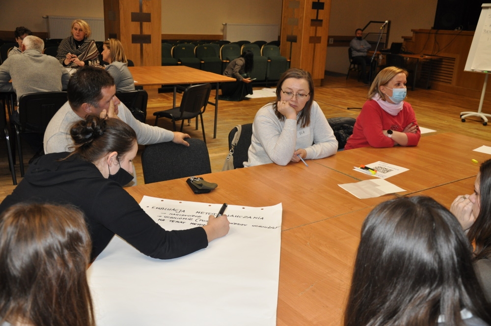 Uczestnicy siedzą przy stole, jedna kobieta notuje na dużej kartce