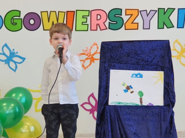Chłopiec w białej koszuli trzyma w ręku mikrofon, po prawej stronie stoi praca plastyczna.