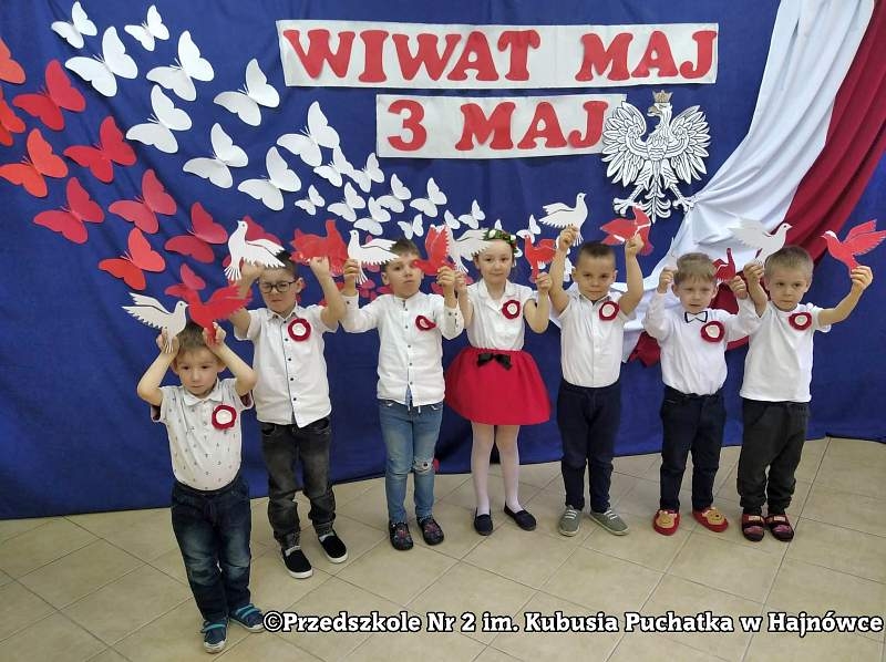 Dzieci ubrane na galowo trzymają biało-czerwone papierowe ptaki.