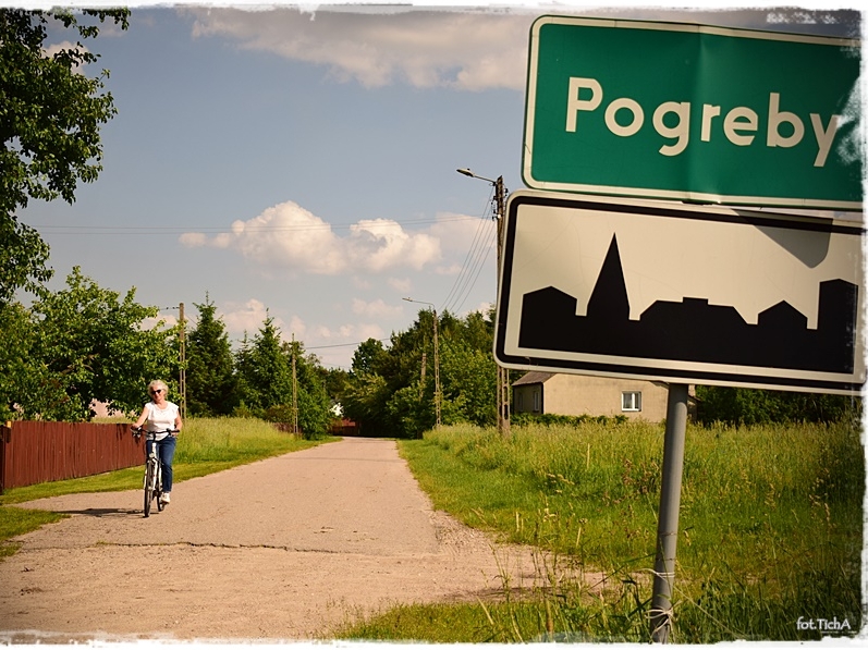 Z prawej strony znak drogowy z nazwą wsi, z lewej po żwirowej drodze jedzie rowerzysta.