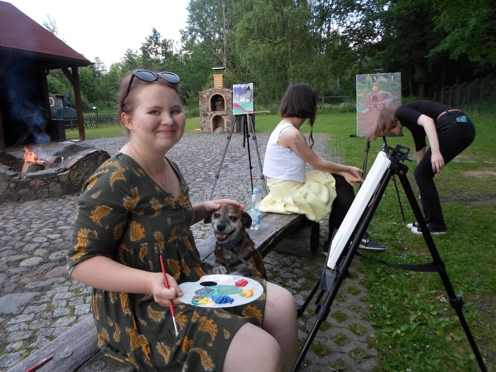 Kobiety stoją i siedzą przy sztalugach, jedna z nich trzyma paletę i pędzel.