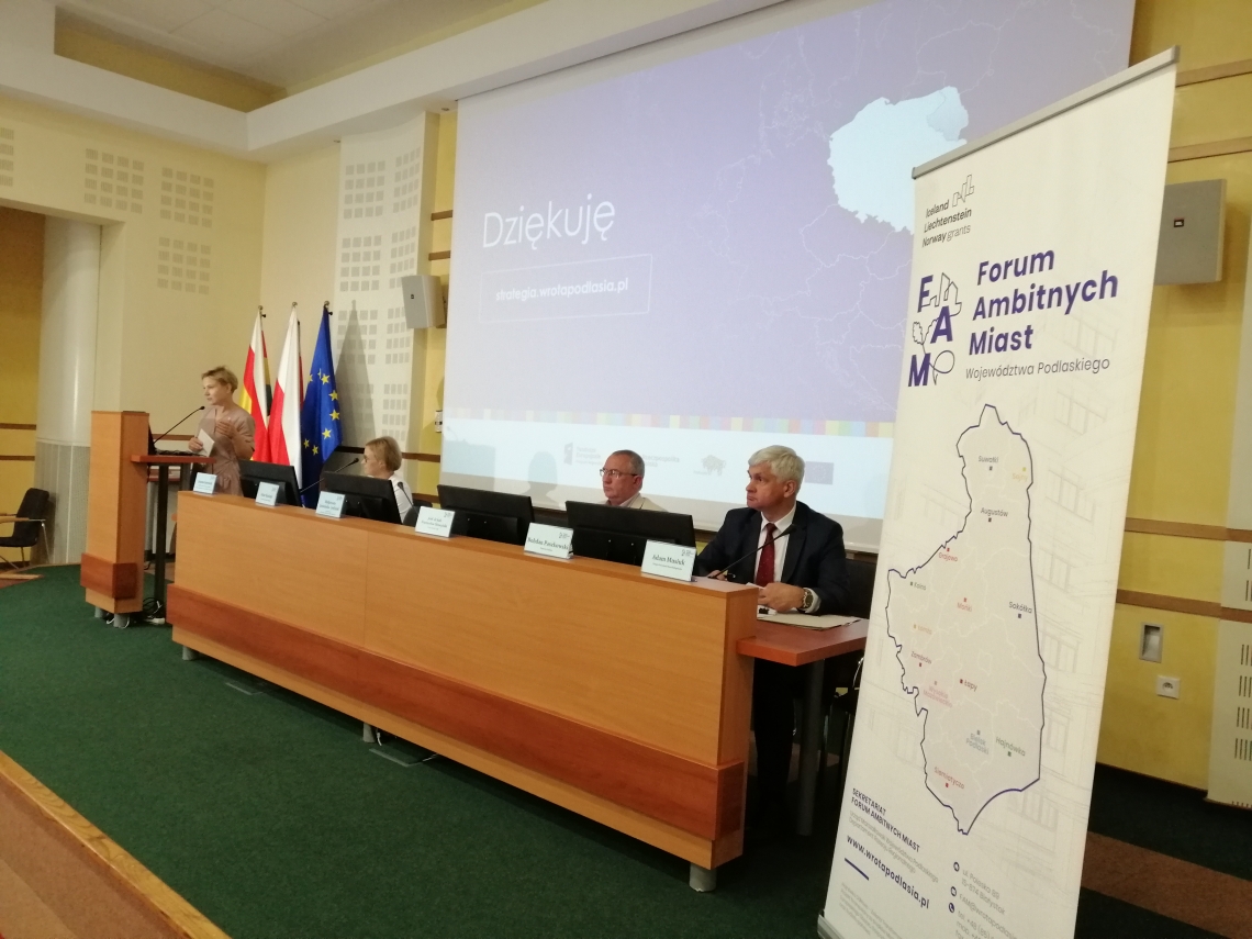 grupa osób siedząca z długim stołem, za nimi wisi duży ekran z wyświetlonym slajdem