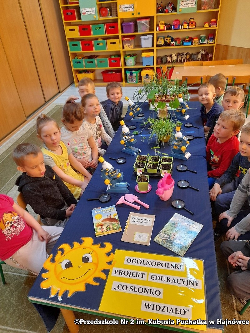 Dzieci dziedzą przy stole z niebieskim obrusem, na stole m. in. lupy.