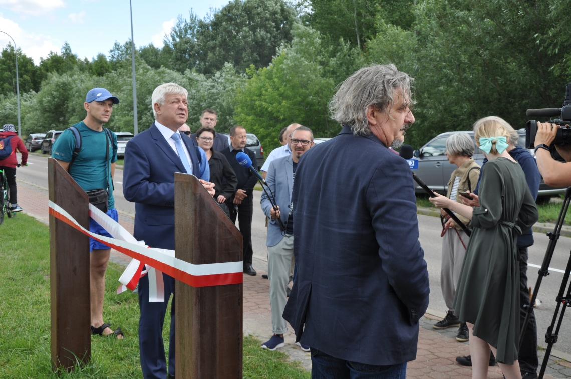 grupa osób stojąca przy dwóch drewnianych słupach medzy którymi zawieszona jest biało-czerwona wstęga