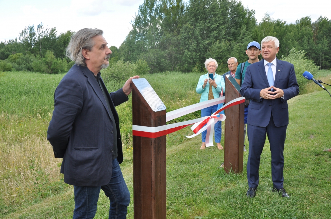 dwóch mężczyzn stoi przy dwóch drewnianych słupach pomiędzy którymi zawieszona jest biało-czerwona wstęga