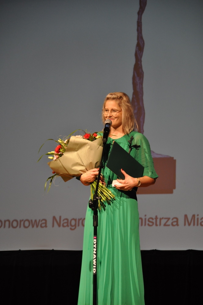Laureatka przemawia do mikrofonu, trzyma bukiet kwiatów i statuetkę.
