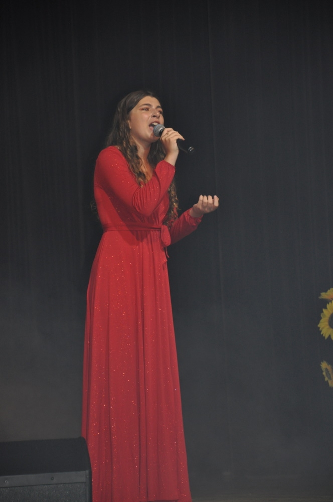Dziewczyna w czerwonej, długiej sukni śpiewa na scenie, w ręku trzyma mikrofon.