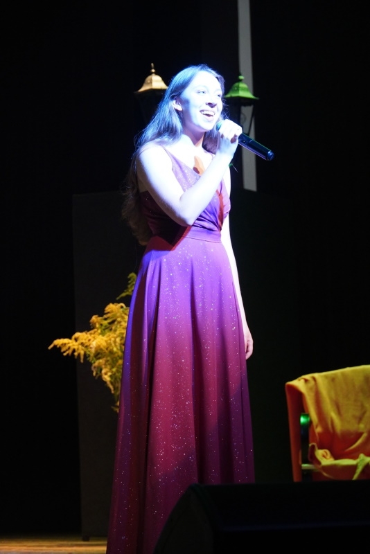 Dziewczyna w czerwonej sukience śpiewa do mikrofonu, który trzyma w ręku.