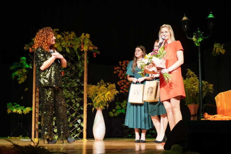 Na zdjęciu stojące cztery kobiety, kobieta w czerwonej sukience trzyma bukiet kwiatów i przemawia do mikrofonu.