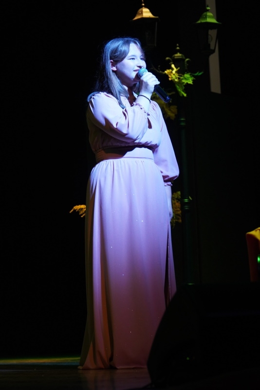 Dziewczyna w jasnej sukience śpiewa do mikrofonu, który trzyma w ręku.