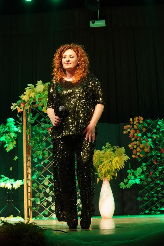 Kobieta w zielonym błyszczącym stroju śpiewa do mikrofonu, który trzyma w ręku.