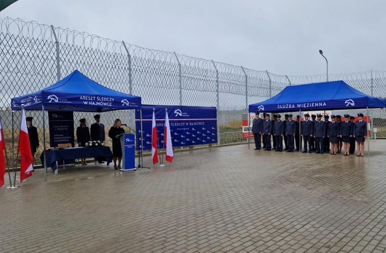 Po lewej i po prawej stronie pod namiotem stoją funkcjonariusze w galowych mundurach, w tle ogrodzenie, deszczowa pogoda.