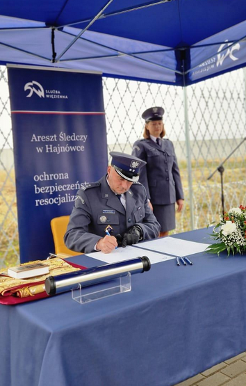 Przy stole siedzi funkcjonariusz podpisujący dokumenty. Z tyłu stoi funkcjonariuszka