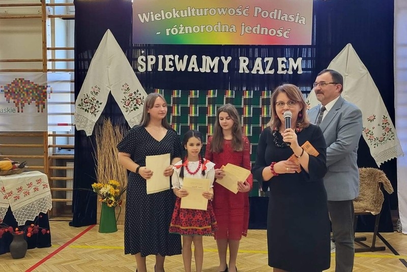 Trzy dziewczynki z dyplomami , mężczyzna oraz przemiawiająca do mikrofonu kobieta. W tle widać tematyczną, regionalną scenografię.