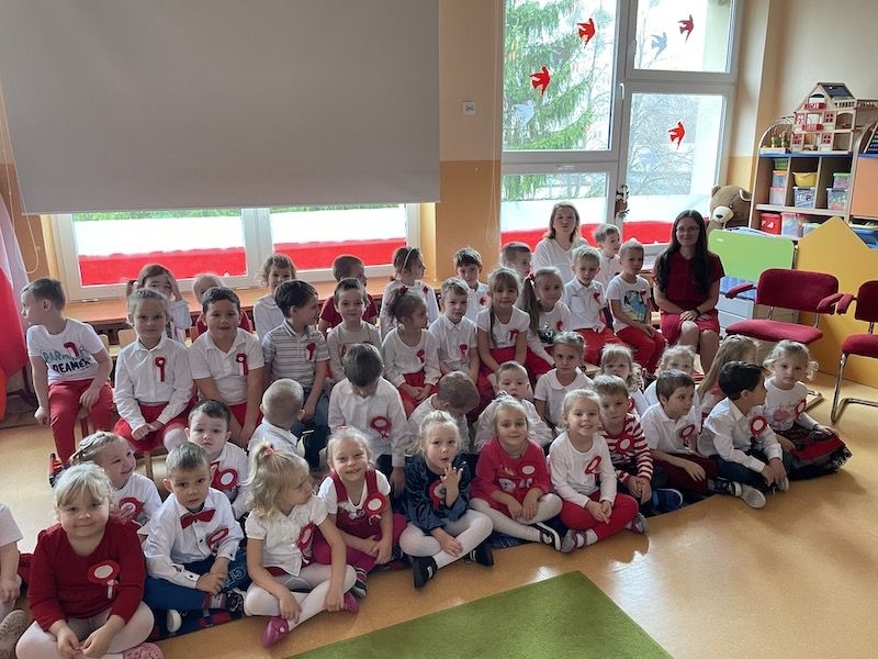 Dzieci siedząc na krzesłach i na podłodze, ubrane są w biało - czerwone stroje.