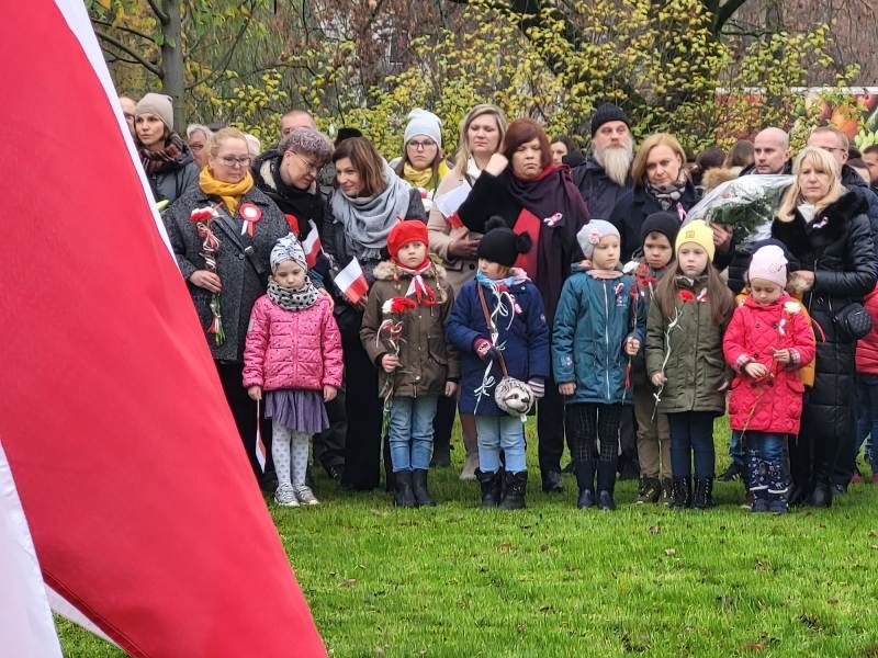 Delegacja dzieci i nauczycieli stojących przed pomnikiem w parku, składających kwiaty.