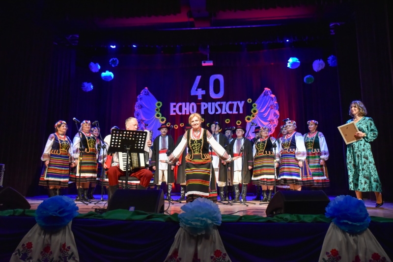 Na scenie stoją kobiety ubrane w kolorowe, ludowe stroje, na krześle siedzi akordeonista.