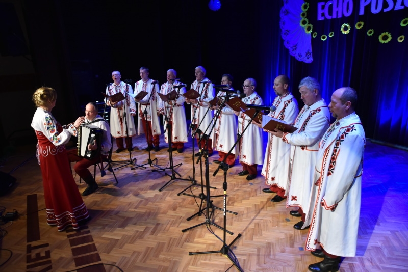 Mężczyźni ubrani w białe stroje stoją na scenie, przed nimi dyrygentka i akordeonista.