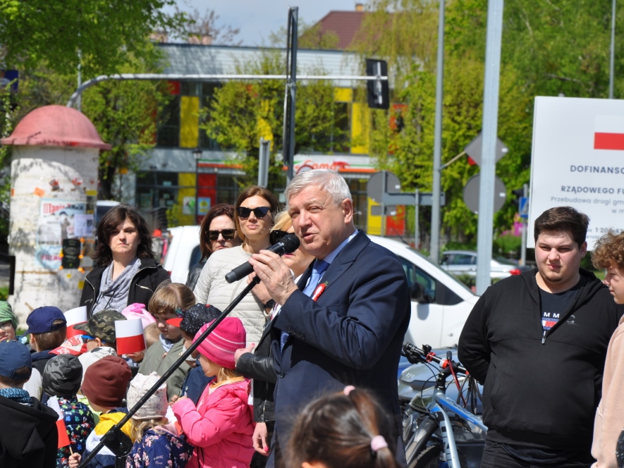 Burmistrza przemawia do mikrofonu, w tle dzieci przedszkolne i mieszkańcy miasta.