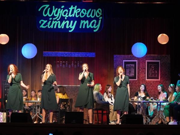 grupa dziewcząt w zielonych sukienkach stoi na pierwszym planie sceny, z tyłu widac inne osoby siedzace przy stolikach. Nad scena wpodświetla się napis "Wyjątkowo zimny maj"