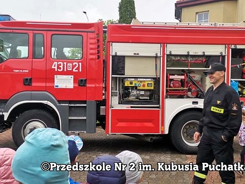 dwóch strażaków pokazuje dzieciom sprzęt mieszczący się w wozie strażackim