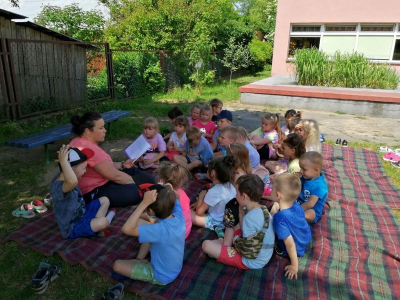Dzieci siedzą i leżą na kocach w ogrodzie przedszkolnym, słuchają czytanych przez panie książek.