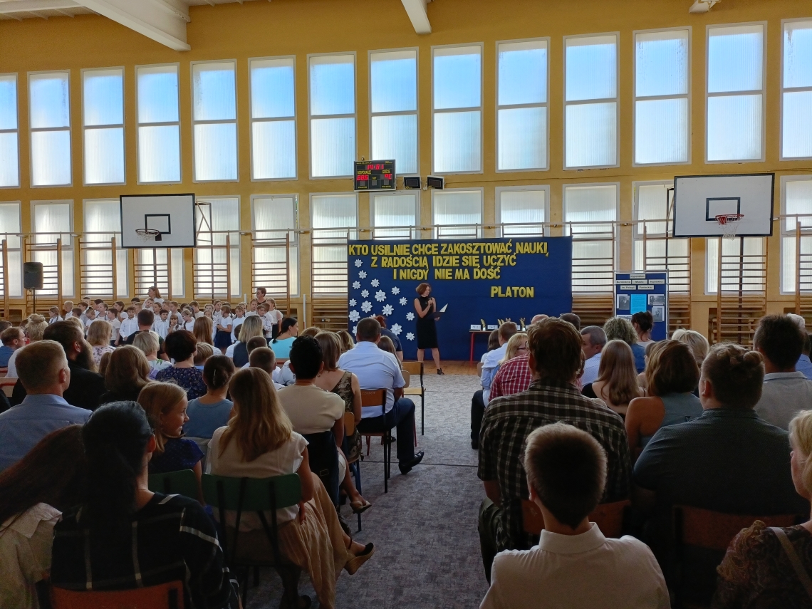 rodzice z dziećmi i nauczyciele zgromadzeni na uroczystości w sali gimnastycznej