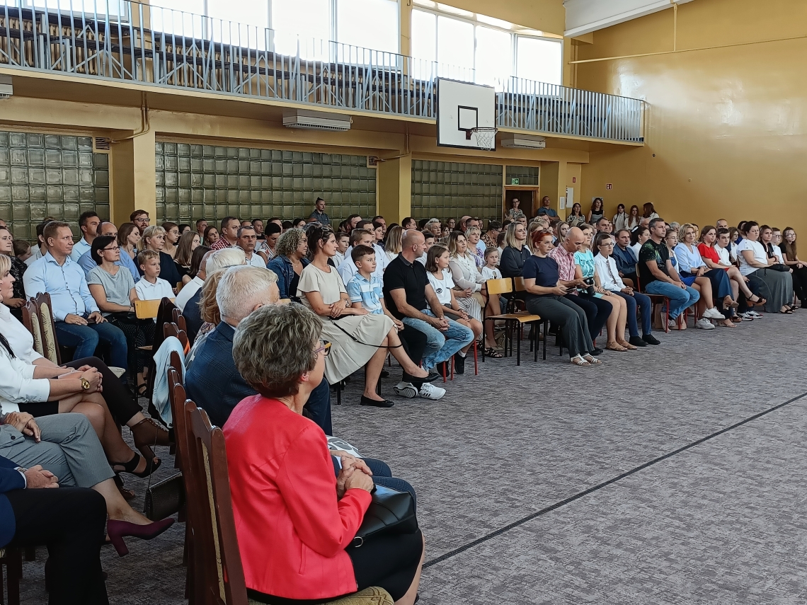 rodzice z dziećmi i nauczyciele zgromadzeni na uroczystości w sali gimnastycznej