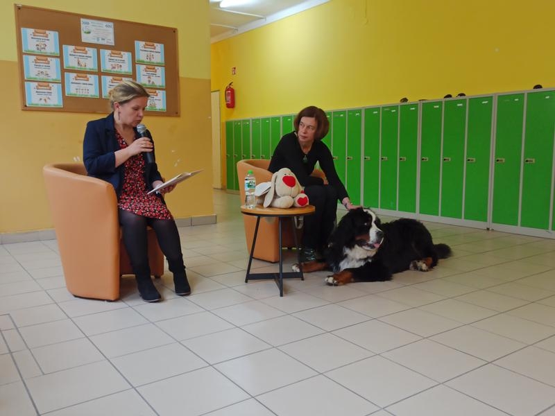 dwie siedzące kobiety. Jedna trzyma książkę, druga głaszczedużego czarnego  psa