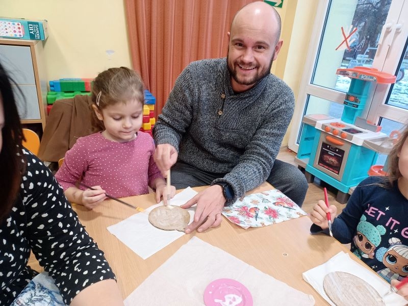 przedszkolaki siedzą przy stołach wraz ze swoimi bliskimi. Wspólnie wykonują ozdoby choinkowe