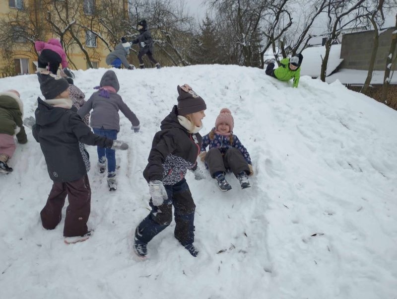 Na podwórku dzieci bawią się na górce, zjeżdżają na pupach, robią anioły na śniegu, biegają po placu.