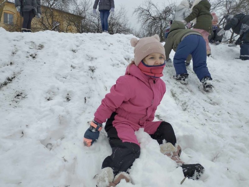 Na podwórku dzieci bawią się na górce, zjeżdżają na pupach, robią anioły na śniegu, biegają po placu.