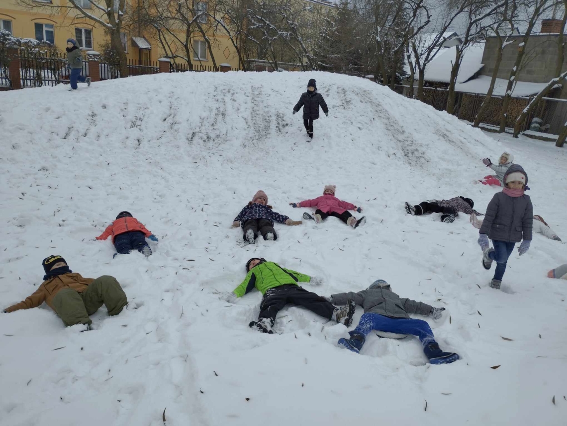 Na podwórku dzieci bawią się na górce, zjeżdżają na pupach, robią anioły na śniegu, biegają po placu.