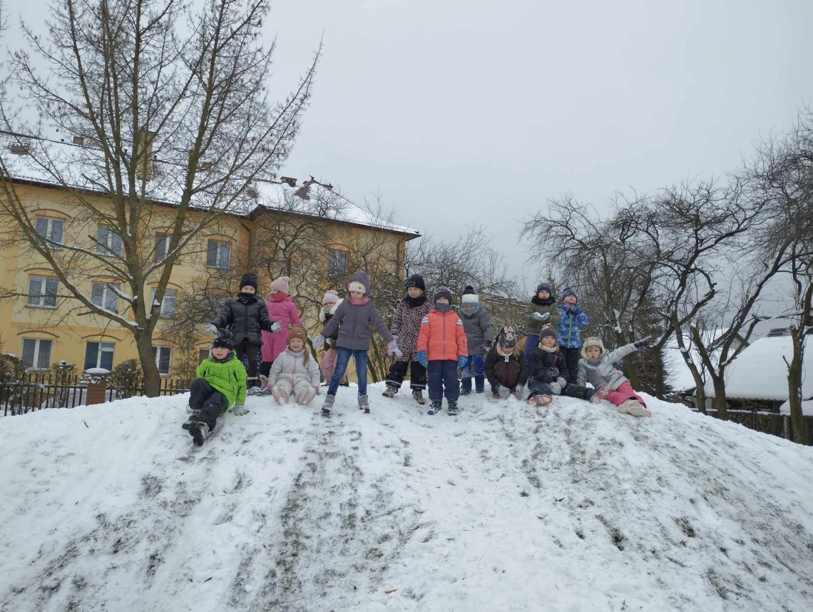 Na podwórku dzieci bawią się na górce, zjeżdżają na pupach, robią anioły na śniegu, biegają po placu.