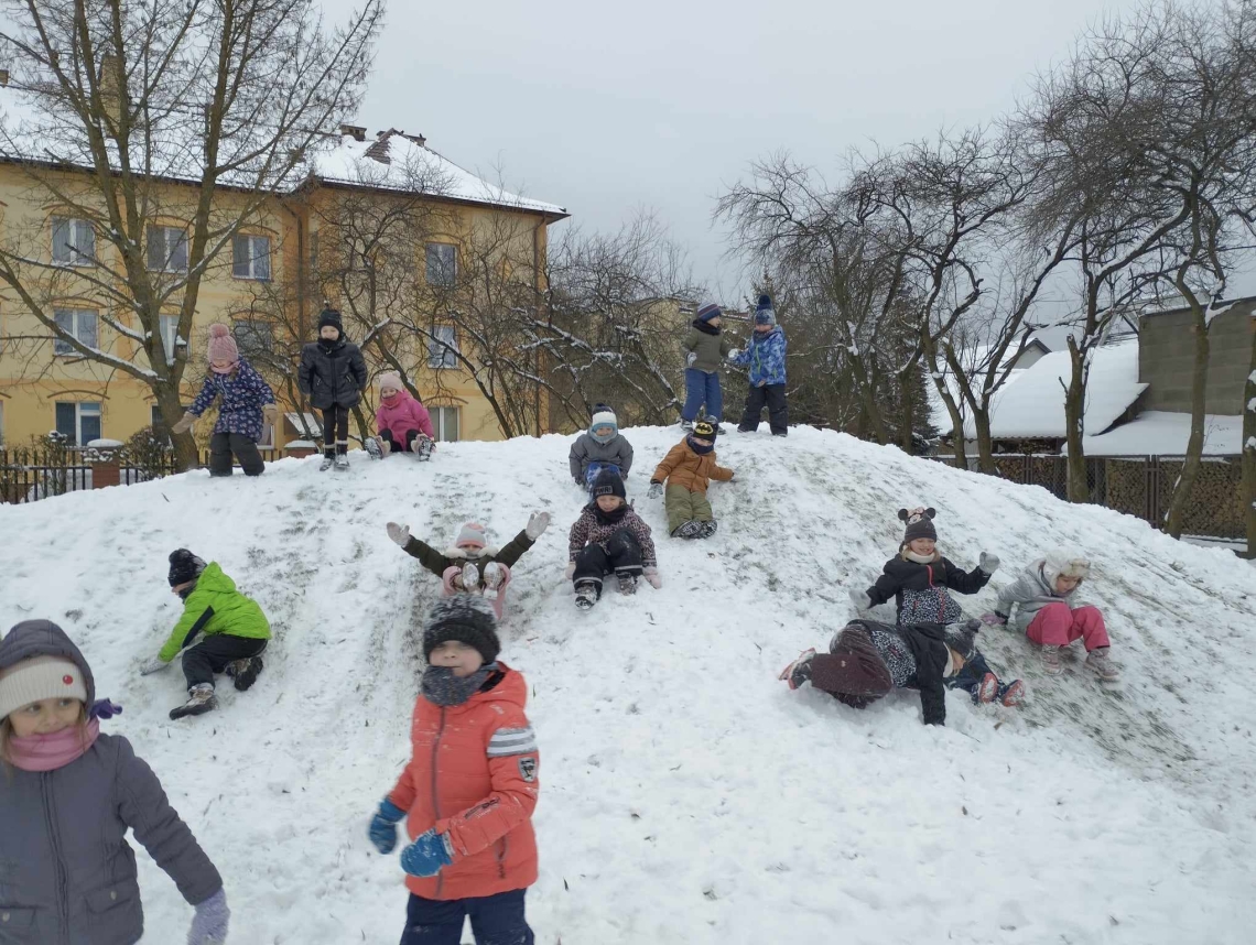 Na podwórku dzieci bawią się na górce, zjeżdżają na pupach, robią anioły na śniegu, biegają po placu.