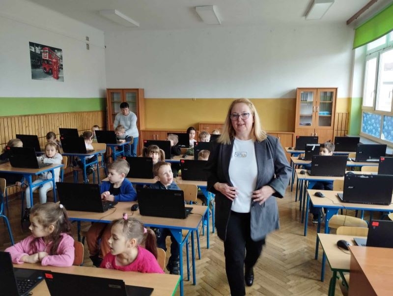 dzieci siedza w ławkach szkolnych. przez śreodek przechodzi kobieta